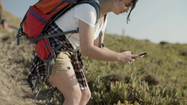 Girl taking photo with her phone while backpacking on sunny day. Dark-haired teenaged girl making photo of flower, exploring nature on hiking adventure. Travel, nature, modern technology concept.