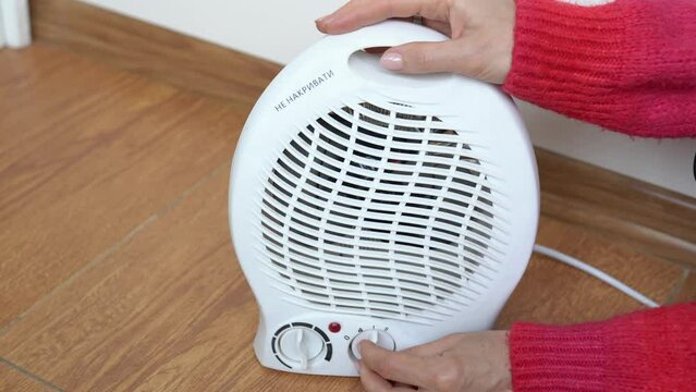 Woman hands in a red sweater adjusting electric fan heater in room and checks with his hand if there is heat. Girl turns on the portable fan heater to heat the room. Autonomous electric space heating