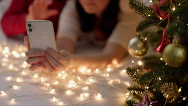 Couple Lies in Bed Uses Smartphone Christmas Pajamas Decorated Christmas Tree Golden Lights Garlands. unrecognizable couple make video call family and friends happy Christmas and New Year concept