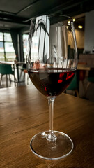 A clean glass with a traditional round shaped glass filled with dark red wine and a thin stem on a wooden table with a blurred restaurant background