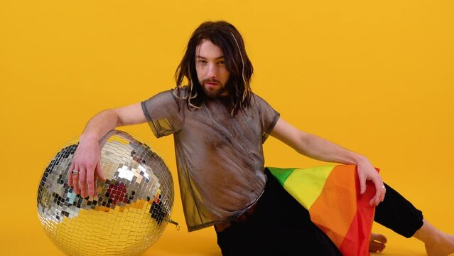 Gay man dressed in mesh t-shirt sits on yellow background with a multicolored flag and silver ball. Concept diversity, transsexual, and freedom