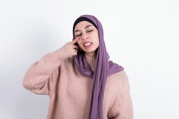 muslim woman wearing hijab and knitted sweater over white background pointing unhappy to pimple on forehead, ugly infection of blackhead. Acne and skin problem