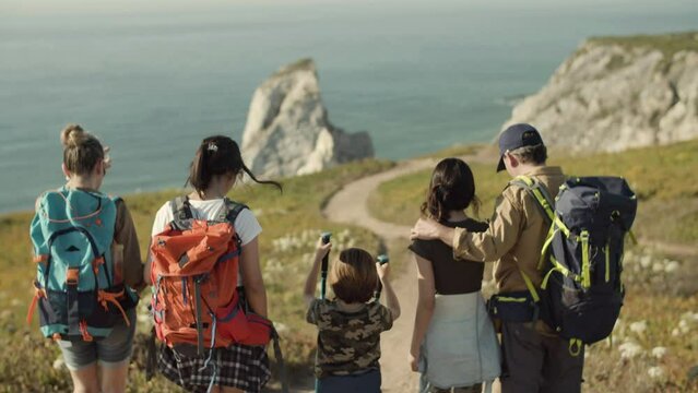 Family with backpacks walking down dirt road towards the sea. Parents and three children on hiking adventure in mountains, spending vacation together. Sea background. Back view. Family, travel concept