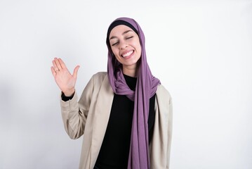 Overjoyed successful young beautiful muslim woman wearing hijab and jacket over white background raises palm and closes eyes in joy being entertained by friends