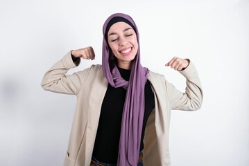 Strong powerful young beautiful muslim woman wearing hijab and jacket over white background toothy smile, raises arms and shows biceps. Look at my muscles!