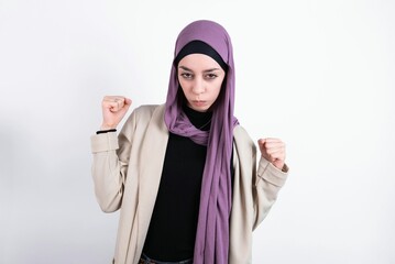 Irritated young beautiful muslim woman wearing hijab and jacket over white background blows cheeks with anger and raises clenched fists expresses rage and aggressive emotions. Furious model