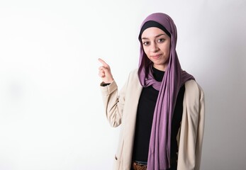 Positive young beautiful muslim woman wearing hijab and jacket over white background with satisfied expression indicates at upper right corner shows good offer suggests to click on link
