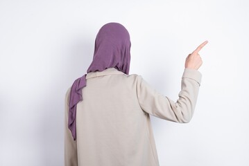 young beautiful muslim woman wearing hijab and jacket over white background pointing to object on copy space, rear view. Turn your back