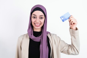 Photo of happy cheerful smiling positive young beautiful muslim woman wearing hijab and jacket over white background recommend credit card