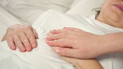 Fototapeta na wymiar Daughter strokes hand of sick mother lying in bed at home. Woman takes care of elderly bedridden mom after difficult surgery showing support closeup