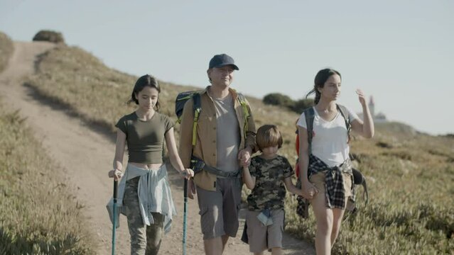 Father and children backpacking together, walking along path, enjoying views around. Daughter using trekking poles while mans holding his sons hand. Slow motion, front view. Family, travel concept.