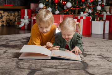 Portrait of little blond boy and sister with book near the Christmas tree. Siblings children on Christmas Eve.