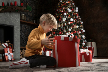 Happy child opens big Christmas gift on Christmas tree background. Joyful boy with Xmas present....