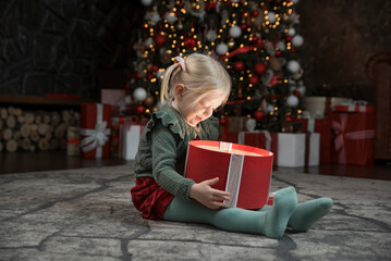 Obraz na płótnie Canvas Happy little girl opening xmas gift near Christmas tree. Gift is shine from inside. Cute blonde girl with gift box.