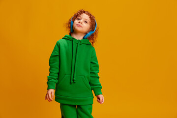 Portrait of cute little girl, child with curly red hair posing in headphones isolated over yellow background