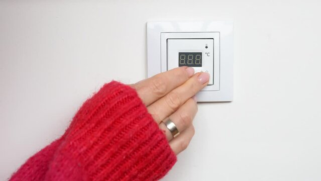 Woman hand in a red sweater setting temperature on underfloor heating control panel. Freezing girl raises the temperature in room by pressing the button on the floor heating thermostat panel on wall