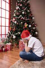The boy sits near the Christmas tree turned away sad waiting for Santa Claus