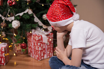 The boy sits near the Christmas tree turned away sad waiting for Santa Claus