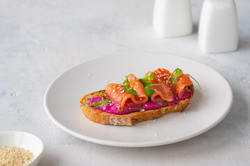Open sandwich with beetroot puree, arugula and salted salmon on a white plate on a light concrete background. Recipes for sandwiches, smorrebrods.