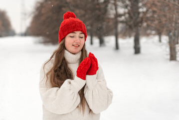 Smiling pretty girl in red knitted hat and mittens and woolen sweater on alley in winter park. Woman outdoor