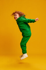 Portrait of cute little girl, child with curly red hair posing, cheerfully jumping isolated over yellow background