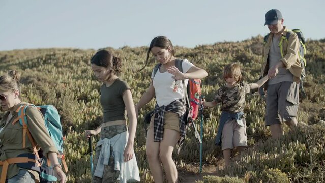 Caucasian family getting down steep slope hiking in mountains. Parents and children backpacking on vacation together, carefully descending hill, using trekking poles. Front view. Family, trip concept