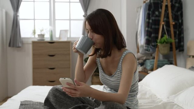 young asian japanese woman in pajama sitting on bed and drinking cup of coffee. lazy girl looking smart phone screen at the sunny morning bedroom. female typing on cellphone and laughing with hot tea