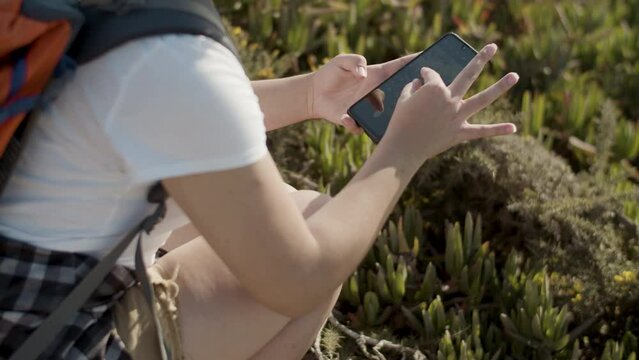 Closeup of Caucasian girl with backpack making photo of flowers. Young woman taking picture with mobile phone while walking along hiking trail. Handheld shot. Nature, travel, modern technology concept