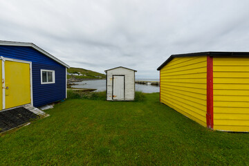 Fototapeta na wymiar Multiple colorful wooden beach huts on vibrant green grass. The small buildings are yellow, blue, red, and white colored. The blue shed has a vibrant double yellow door with a ramp up the door.