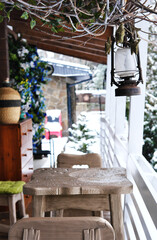 Terrace of a wooden house overlooking the winter forest. There are a wooden table, chairs and a lantern on the terrace.