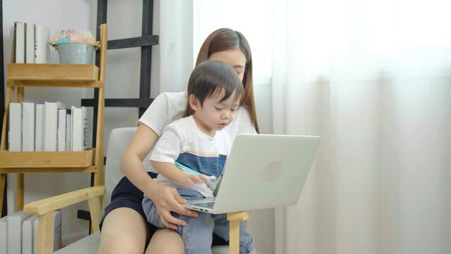 Young asian mother her little adorable son using laptop looking at computer screen enjoying watching funny social media video doing online shopping relaxing on sofa at home together