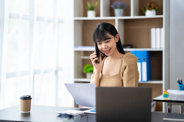 Happy young asian woman talking on the mobile phone and smiling while sitting at her working place in office.
