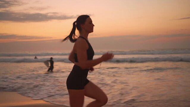 Athletic woman with waving hair running on beach at dramatic sun background slow motion. Sportswoman enjoying outdoor physical activity on seaside at sunset side view.