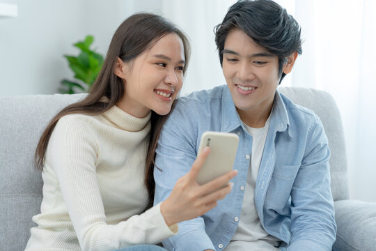 Asian couple smiling and happily use smartphone to online shopping. Husband ready to pay birthday present for beautiful wife. Find accommodation online for honeymoon, App online, book flight tickets.