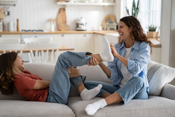 Laughing mom and daughter have fun as best friends spending time together for happy family life. Joyful Woman in casual clothes and school age girl playing tickling sits on sofa. Love in the family - obrazy, fototapety, plakaty
