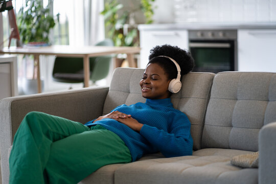 Resting Casual African American Girl In Headphones With Slight Smile And Closed Eyes On Couch. Positive Black Woman Falls Asleep Lying In Living Room Listening To New Music Album Of Favorite Band