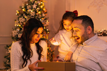 Happy family with surprising emotion while opening a gift box there is a christmas tree, fireplace and candles on the background.