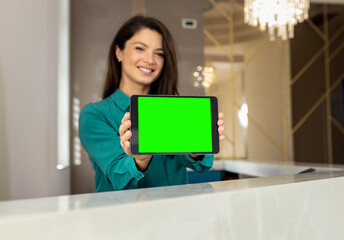 Portrait of female receptionist showing tablet with green screen towards the camera at reception...