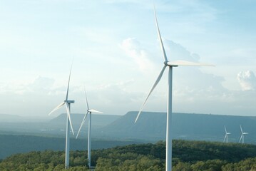 Field in the countryside with tall wind turbines sticking out of thick forests under a bright sky