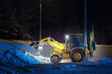 Backhoe loader cleans snow on parking lot outside the city, night snow removal work. Snow clearing,...