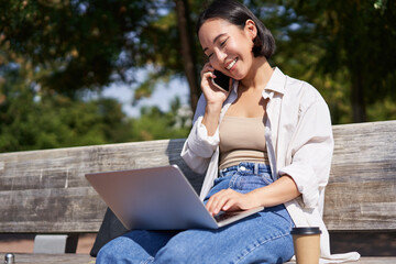Young asian girl talking on mobile phone, using laptop, working online from park, sitting on bench on sunny day with computer