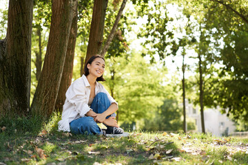 Portrait of beautiful asian woman resting near tree, relaxing in park, smiling and looking happy