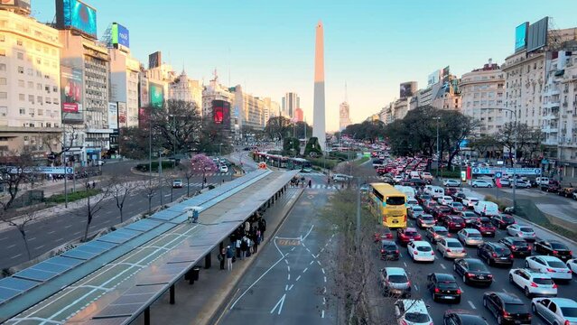 Cinematic aerial dolly in shot fly along 9 de julio avenue towards obelisco historical monument, icon of Buenos Aires city at Plaza de la Rep&uacute;blica downtown, capturing busy traffics at rush hour.