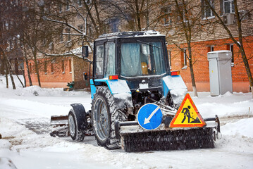 Tractor with brush and scoop cleaning snow at city street during heavy snowfall at winter day. Tractor clearing snowy streets with snow plow and ratary brush. Vehicle sweeps snow on pedestrian road
