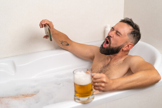 Bearded man watching football on smartphone while drinking a beer in bath with soapsuds at home