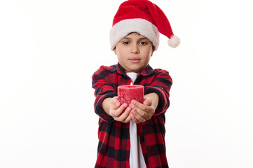 Adorable Caucasian preteen boy wearing Santa hat, holding a lit red candle to the camera, posing against a white background. Copy ad space. Merry Christmas and Happy New Year concept. Winter holidays
