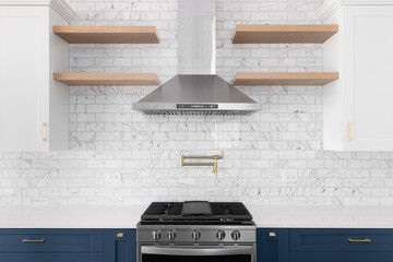 A kitchen detail shot with astainless steel hood, marble subway tile backsplash, white and blue cabinets, and white marble countertops.