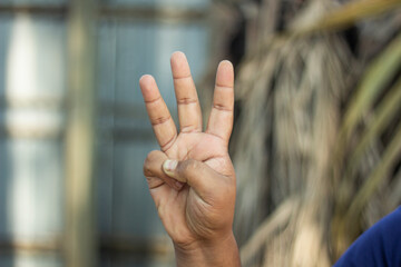 A hand has two fingers folded out of five fingers and the remaining three fingers are pointing upwards and the background is blurred.