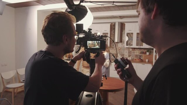 Tracking of cropped cameraman filming advertising video on professional camera, using dollying in technique, two female and male actors visible on screen