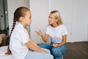 Upset pretty little girl feeling sad sitting side to angry strict blonde mother scolding lecturing difficult kid for bad behavior at home. Mad mom arguing shouting at stubborn child preteen daughter.